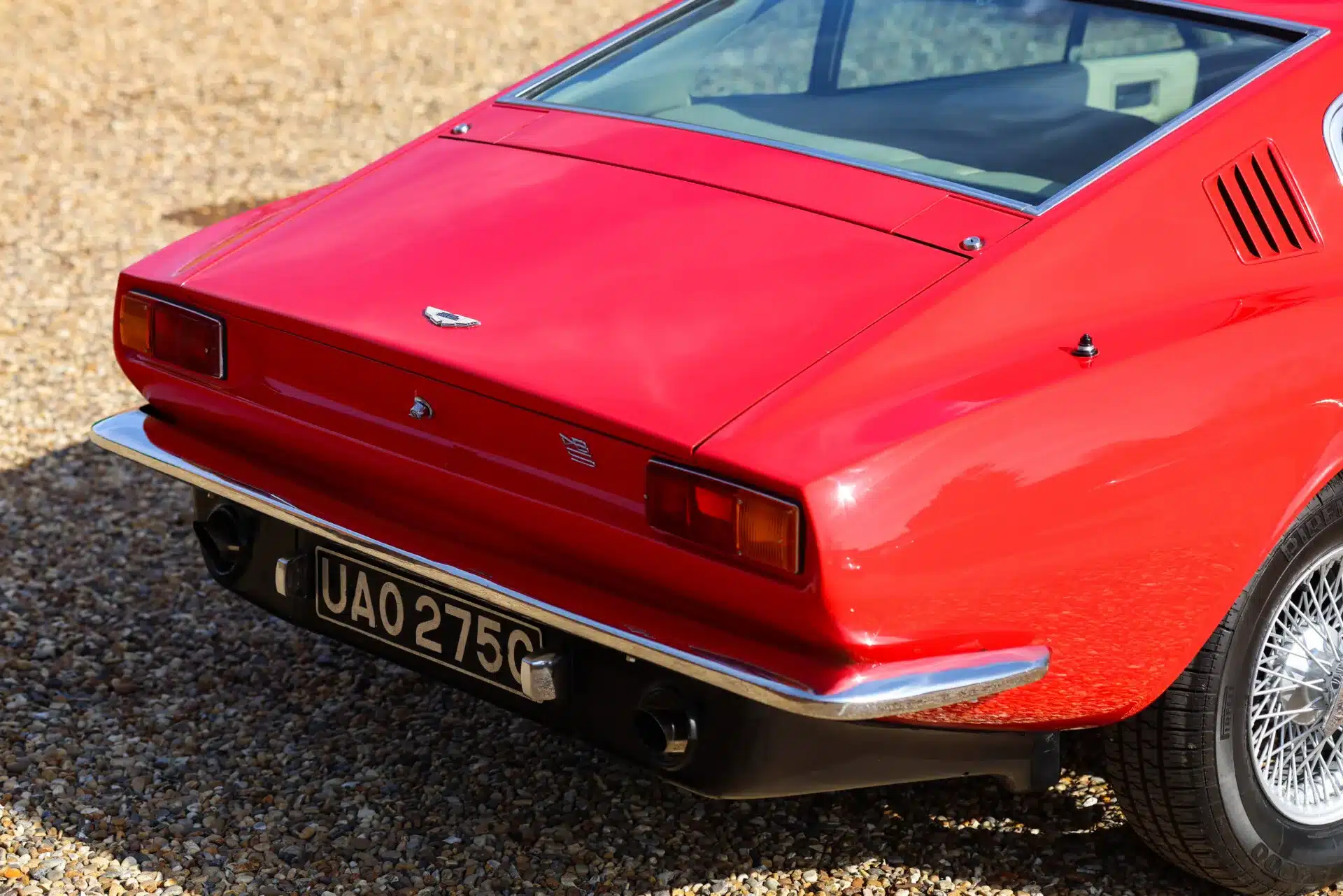 Heckansicht eines roten Aston Martin DBS 1969 mit beigem Innenraum und schwarzem Kennzeichen auf Kiesuntergrund.