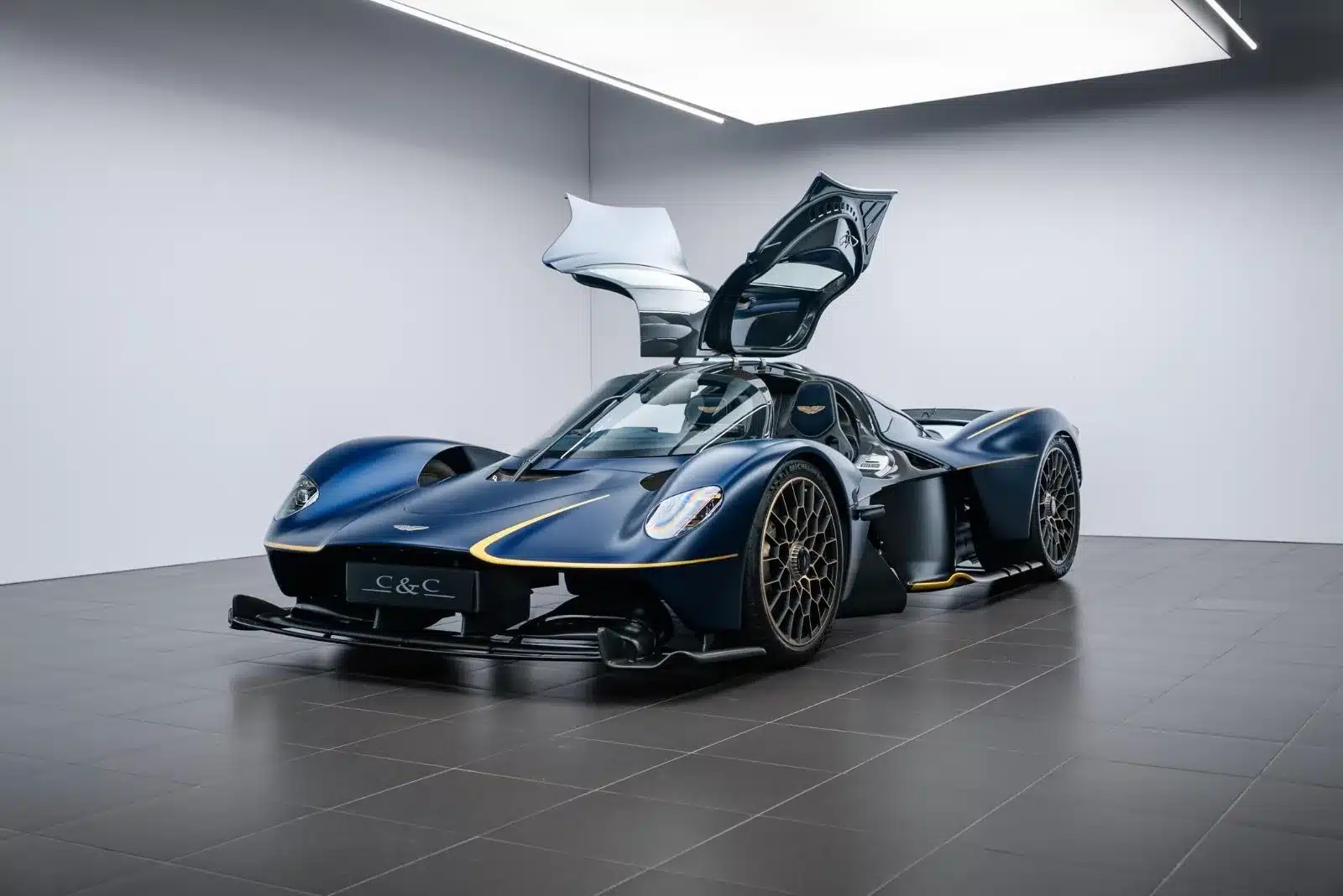 Aston Martin Valkyrie, blauer Supersportwagen mit geöffneten Flügeltüren, in einem hellen Showroom von vorne schräg fotografiert.