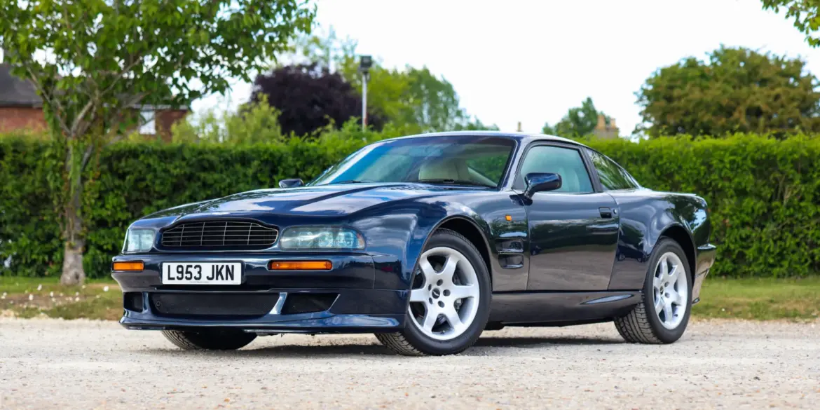 Aston Martin Vantage V550 in dunkelblauer Metallic-Lackierung, schräg von vorne fotografiert, auf einem Kiesplatz vor grüner Hecke.