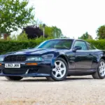 Aston Martin Vantage V550 in dunkelblauer Metallic-Lackierung, schräg von vorne fotografiert, auf einem Kiesplatz vor grüner Hecke.