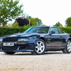 Aston Martin Vantage V550 in dunkelblauer Metallic-Lackierung, schräg von vorne fotografiert, auf einem Kiesplatz vor grüner Hecke.