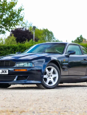 Aston Martin Vantage V550 in dunkelblauer Metallic-Lackierung, schräg von vorne fotografiert, auf einem Kiesplatz vor grüner Hecke.