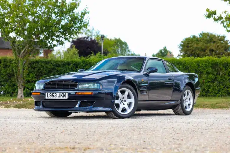 Aston Martin Vantage V550 in dunkelblauer Metallic-Lackierung, schräg von vorne fotografiert, auf einem Kiesplatz vor grüner Hecke.