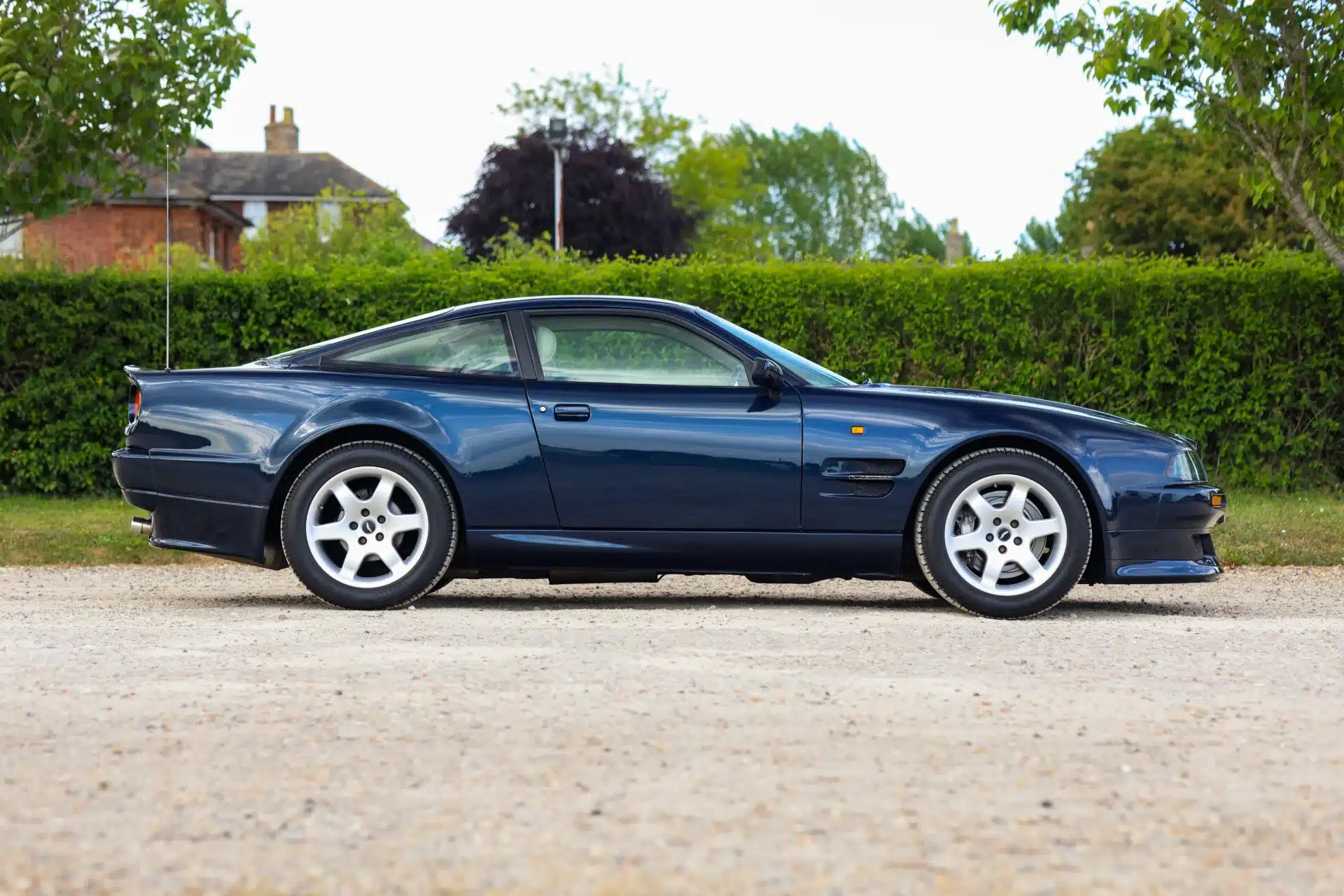 Aston Martin Vantage V550 Baujahr 1994 in Royal Blue metallic, Seitenansicht, geparkt auf Kies mit grüner Hecke im Hintergrund.