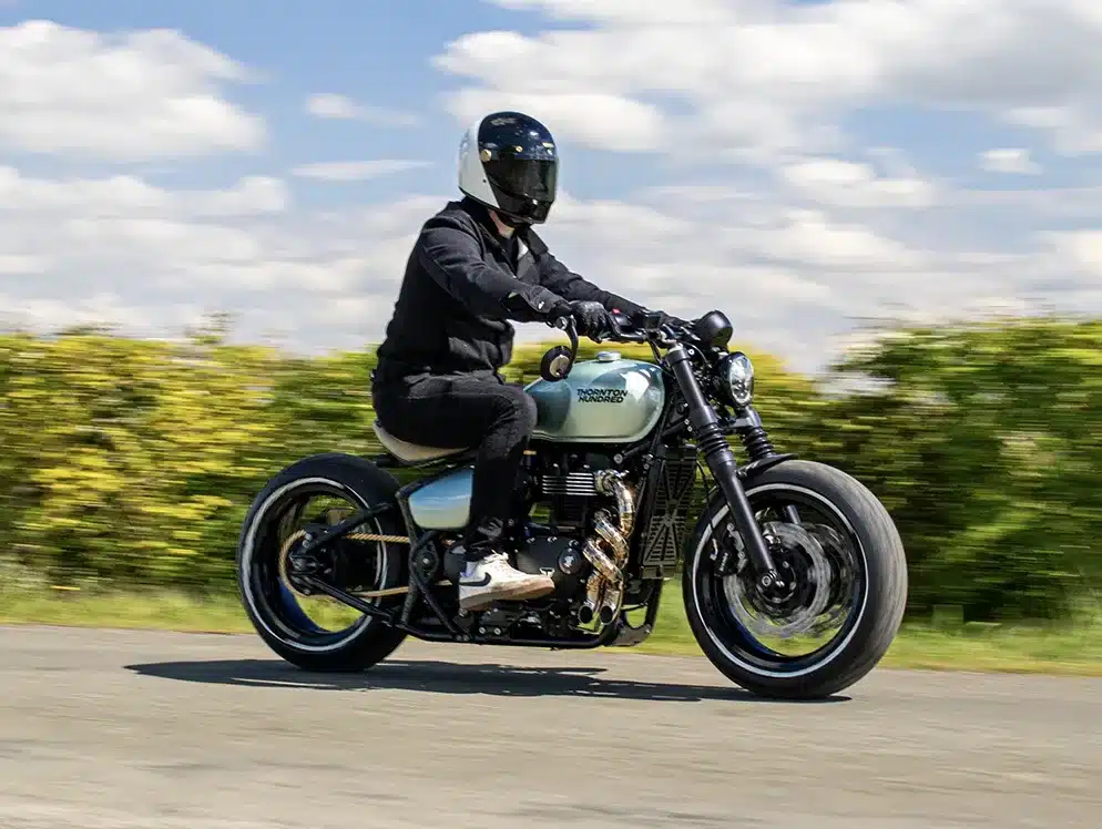 Ein Fahrer mit Helm fährt eine handgefertigte Bespoke Bobber auf einer Landstraße, im Hintergrund Büsche und blauer Himmel mit Wolken.