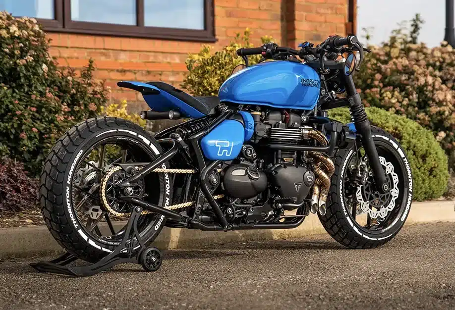 Blaues Custom-Motorrad (Bespoke Bobber) mit handgefertigten Ausstattungsdetails, breiten Reifen und individueller Auspuffanlage auf einem Parkplatz.