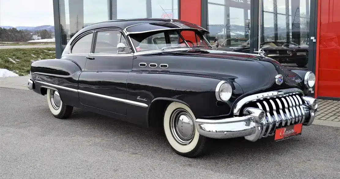 Dunkelblauer Buick Special Jet Back Sedanette von 1950 mit originalem Frontgrill und weißwandbereiften Rädern, seitlich vor einem Autohaus fotografiert.