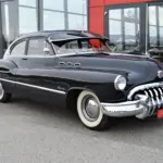 Dunkelblauer Buick Special Jet Back Sedanette von 1950 mit originalem Frontgrill und weißwandbereiften Rädern, seitlich vor einem Autohaus fotografiert.