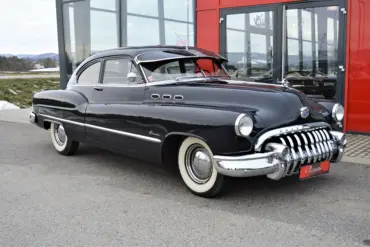 Dunkelblauer Buick Special Jet Back Sedanette von 1950 mit originalem Frontgrill und weißwandbereiften Rädern, seitlich vor einem Autohaus fotografiert.
