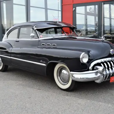Dunkelblauer Buick Special Jet Back Sedanette von 1950 mit originalem Frontgrill und weißwandbereiften Rädern, seitlich vor einem Autohaus fotografiert.