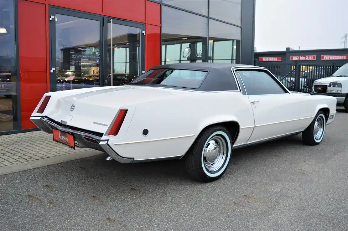 Weißes Cadillac Eldorado Coupe von 1969 mit schwarzem Vinyl-Halo-Dach, fotografiert schräg von hinten rechts vor einem Autohaus.