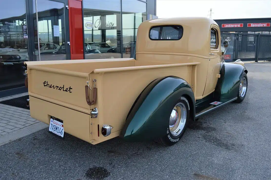 Chevrolet 3100 Pick-up V8 Kompressor Hot Rod von hinten, cremefarben mit grünen Kotflügeln, auf einem Parkplatz vor einer Werkstatt.