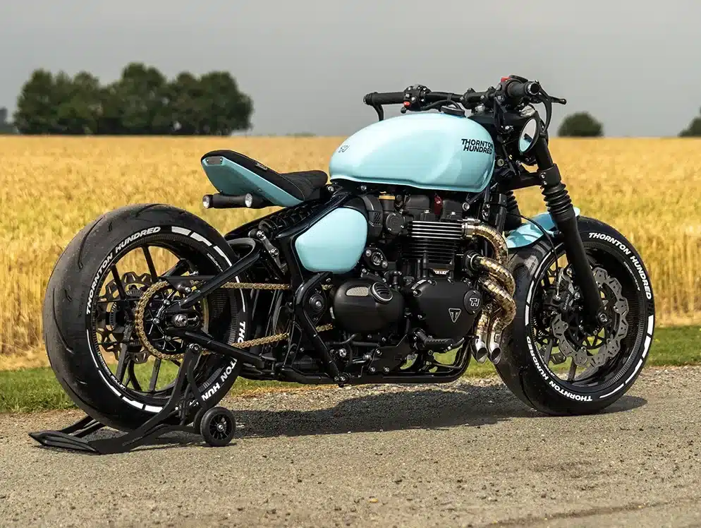Seitenansicht einer hellblauen Thornton Hundred City Bobber mit V4-Carbonrädern und auffälligem Auspuff auf einer Landstraße vor einem Getreidefeld.