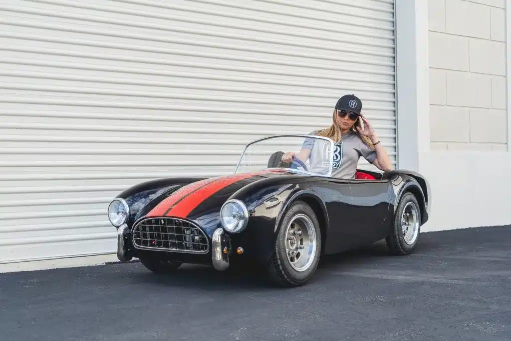 Cobra 289 Junior Kinderauto in Schwarz mit roten Streifen und erwachsener Person am Steuer, abgestellt vor Rolltor.