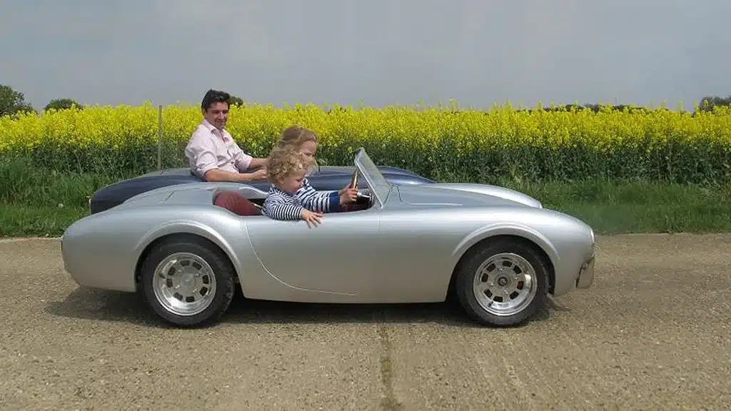 Silberner halbmaßstäblicher Kinder-Roadster Cobra 289 Junior mit Kind und Erwachsenem auf einer Landstraße