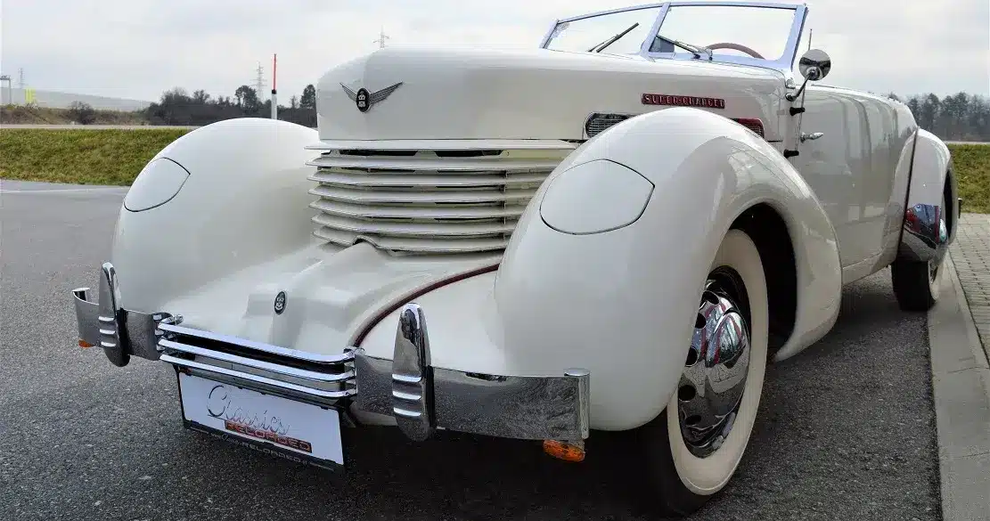 Weißer Cord 812 Supercharged Convertible Phaeton von 1937 mit markantem Kühlergrill und versenkbaren Scheinwerfern auf einer Straße.