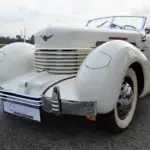 Weißer Cord 812 Supercharged Convertible Phaeton von 1937 mit markantem Kühlergrill und versenkbaren Scheinwerfern auf einer Straße.