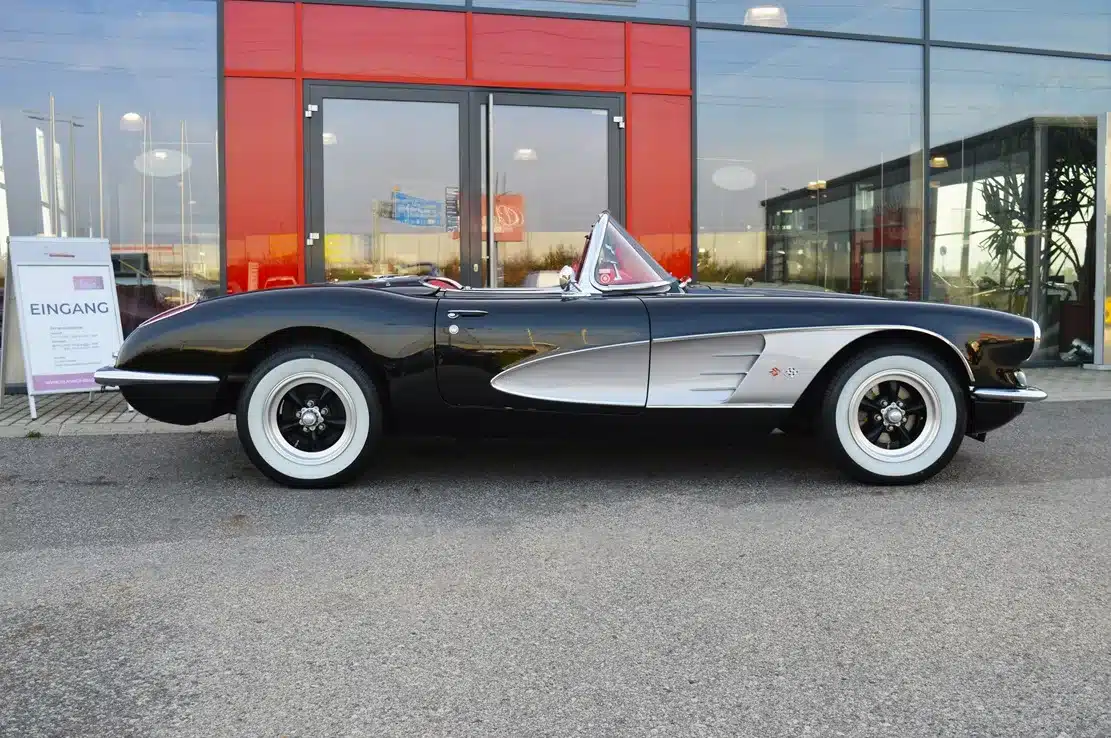 Schwarze Chevrolet Corvette C1 Resto-Mod LT1 Cabrio von 1959 mit weißen Akzenten und Sonnenland-Verdeck, seitliche Ansicht vor gläserner Fassade