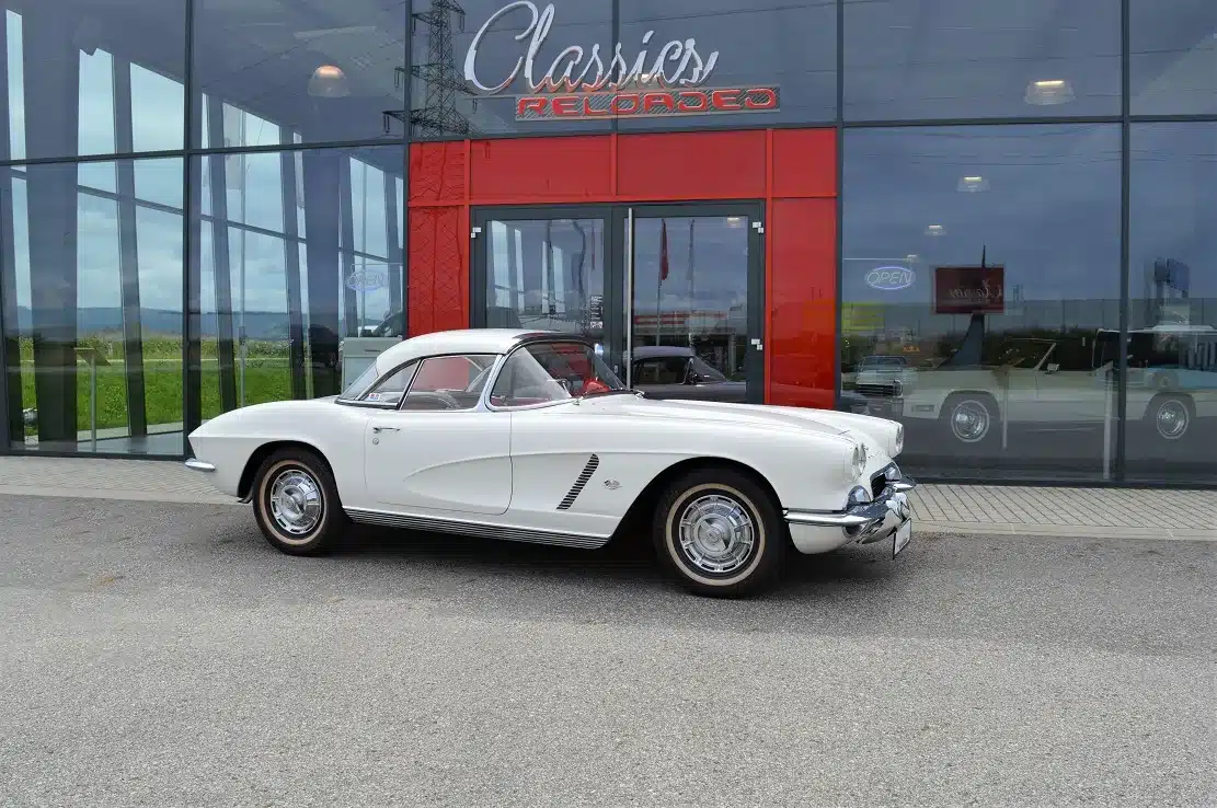 Weiße Corvette C1 Roadster 327 Dual Top Baujahr 1962 mit Hardtop, vor Glasfassade eines Autohauses geparkt.