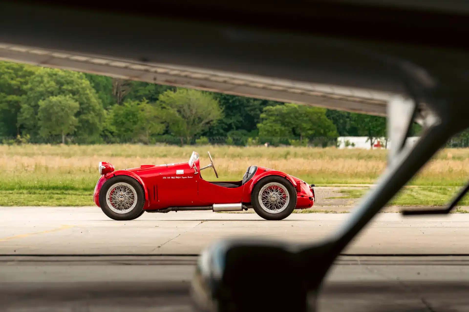 Dagrada Giannini Fiat 750 Sport, Baujahr 1949, rotes Leichtbau-Rennfahrzeug mit mittigem Frontscheinwerfer, seitlich fotografiert auf einem Flugplatz, im Hintergrund grüne Bäume und Wiese.