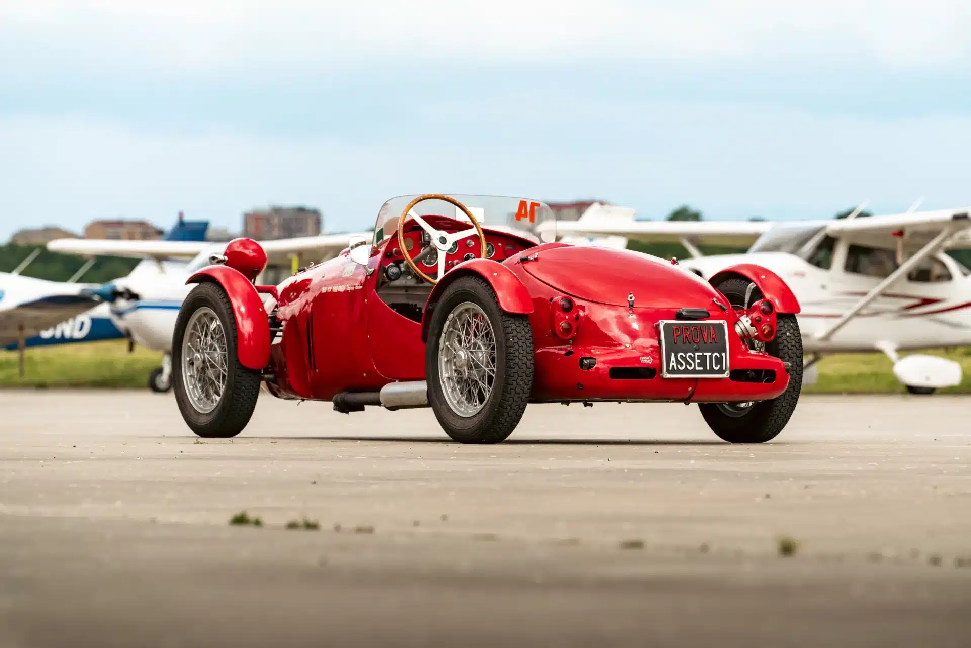 Roter Dagrada Giannini Fiat 750 Sport von 1949 mit mittigem Einzelscheinwerfer und Rennhistorie, geparkt auf Asphalt vor Kleinflugzeugen.