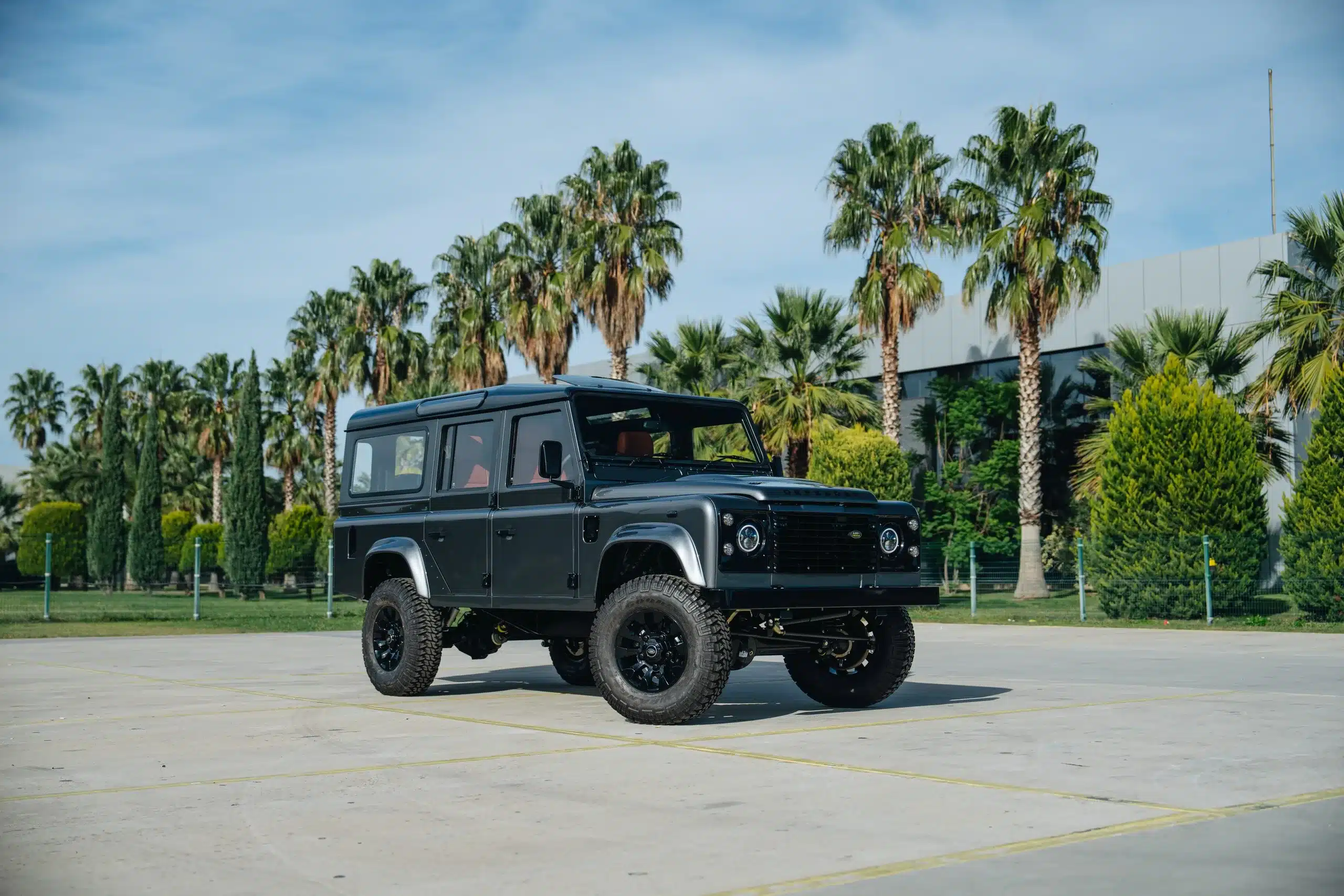 Seitliche Frontansicht eines Land Rover Defender 110 in Corris Grey, parkend auf einem Platz mit Palmen im Hintergrund.