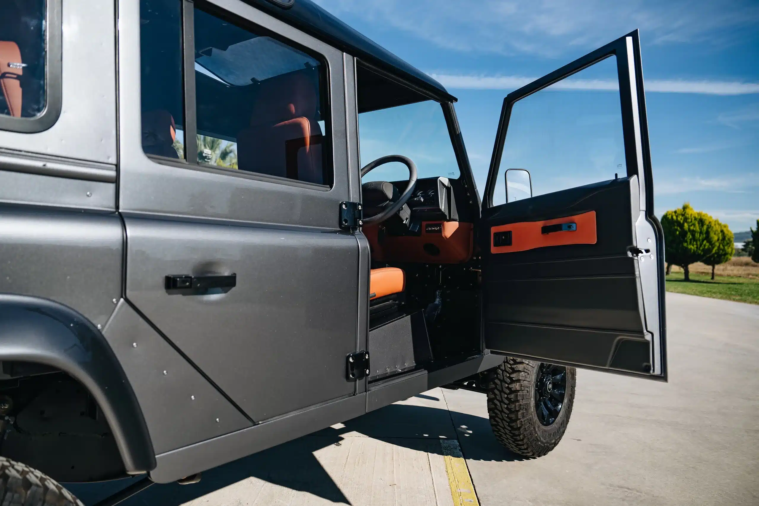 Geöffnete Fahrertür eines Land Rover Defender 110 in Metallic-Grau, Innenausstattung in Orange, Fotografie im Freien bei Sonnenschein.