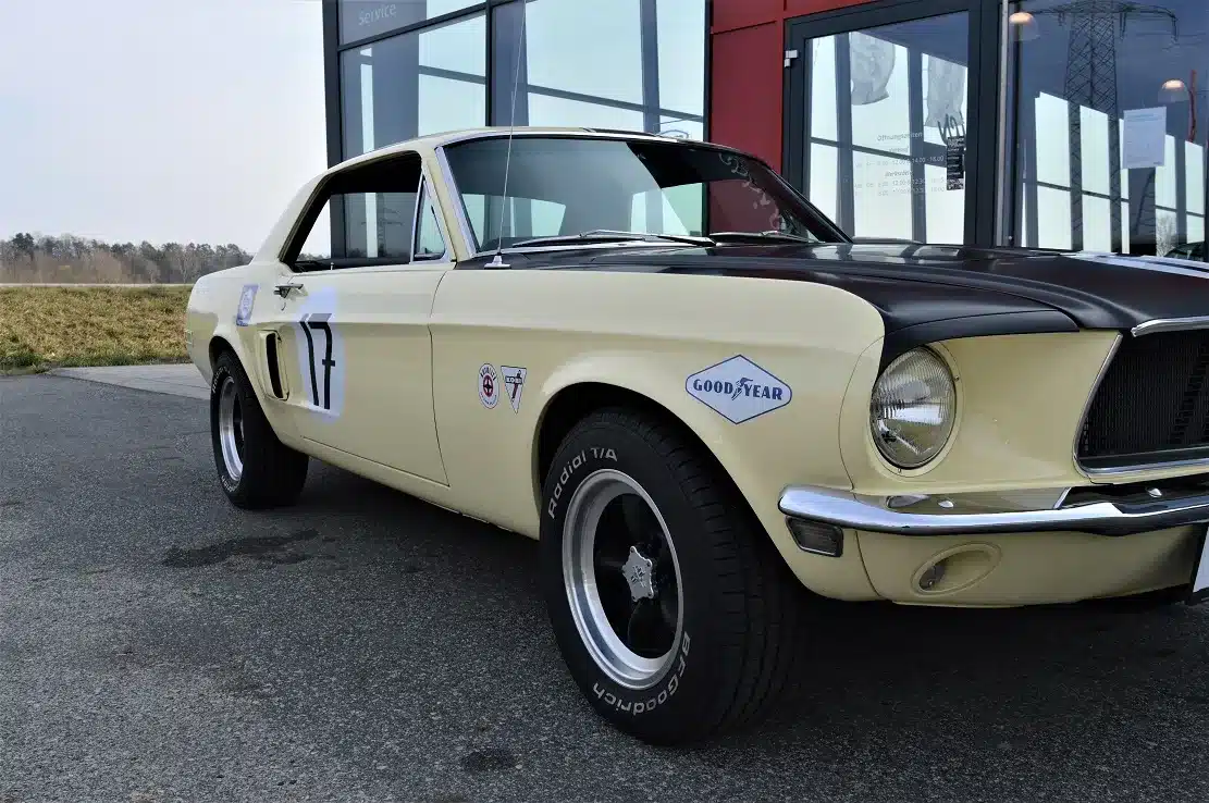 Ford Mustang Shelby Terlingua 302 von 1968 in Meadowlark Yellow mit schwarzer Motorhaube, Startnummer und Rennsport-Beschriftung, geparkt vor moderner Kulisse.
