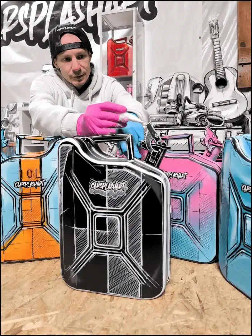 Handbemalter 10-Liter-Kanister in Schwarz, individuell gestaltet mit Acrylfarbe, umgeben von weiteren farbigen Kanistern und einem Künstler bei der Arbeit.