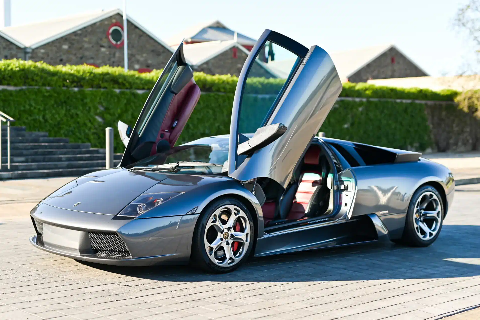 Lamborghini Murciélago 2004, Grigio Avlon, mit geöffneten Scherentüren und rotem Lederinterieur, schräg von vorne fotografiert.