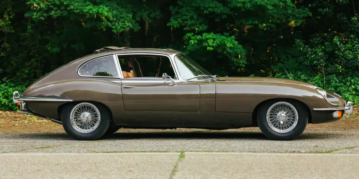 Jaguar E-Type Series 2 2+2 Coupé, Baujahr 1970, braune Lackierung, Seitenansicht, mit geöffnetem Schiebedach, vor grüner Waldkulisse.