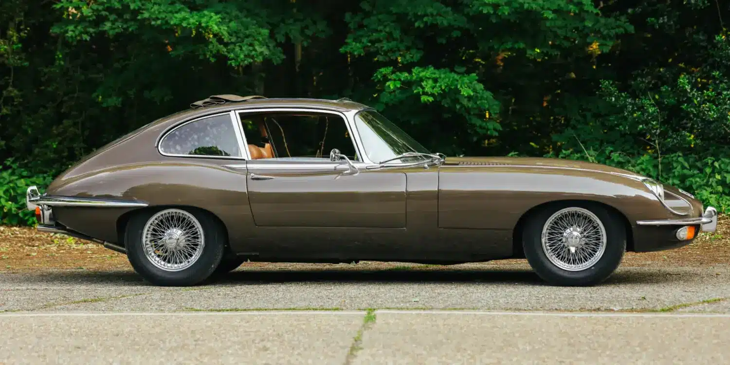 Jaguar E-Type Series 2 2+2 Coupé, Baujahr 1970, braune Lackierung, Seitenansicht, mit geöffnetem Schiebedach, vor grüner Waldkulisse.