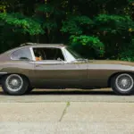 Jaguar E-Type Series 2 2+2 Coupé, Baujahr 1970, braune Lackierung, Seitenansicht, mit geöffnetem Schiebedach, vor grüner Waldkulisse.