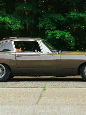Jaguar E-Type Series 2 2+2 Coupé, Baujahr 1970, braune Lackierung, Seitenansicht, mit geöffnetem Schiebedach, vor grüner Waldkulisse.