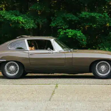 Jaguar E-Type Series 2 2+2 Coupé, Baujahr 1970, braune Lackierung, Seitenansicht, mit geöffnetem Schiebedach, vor grüner Waldkulisse.