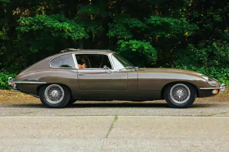 Jaguar E-Type Series 2 2+2 Coupé, Baujahr 1970, braune Lackierung, Seitenansicht, mit geöffnetem Schiebedach, vor grüner Waldkulisse.