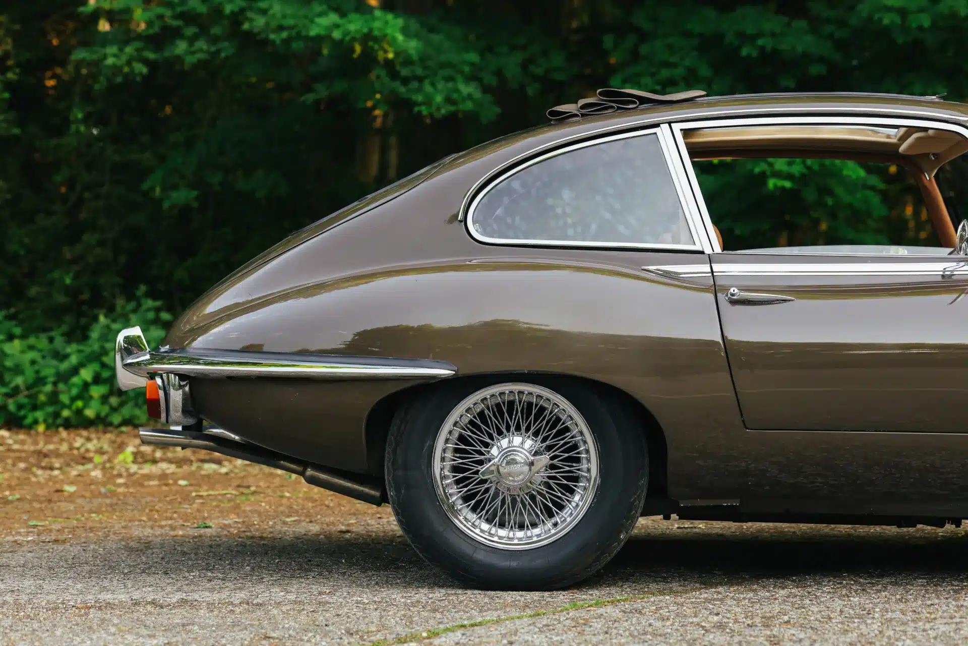 Seitenansicht des hinteren Bereichs eines braunen Jaguar E-Type Series 2 2+2 Coupés von 1970 mit Drahtspeichenrad vor grünem Hintergrund.