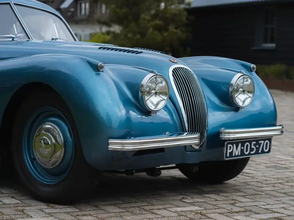 Frontpartie eines blauen Jaguar XK 120 FHC 1951 mit ovalem Kühlergrill und runden Scheinwerfern, aufgenommen im Stand auf Pflastersteinen.