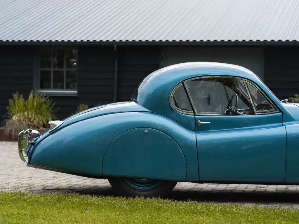 Seitenansicht eines blauen Jaguar XK 120 FHC 1951, parkend auf einer Einfahrt mit Rasen im Vordergrund.