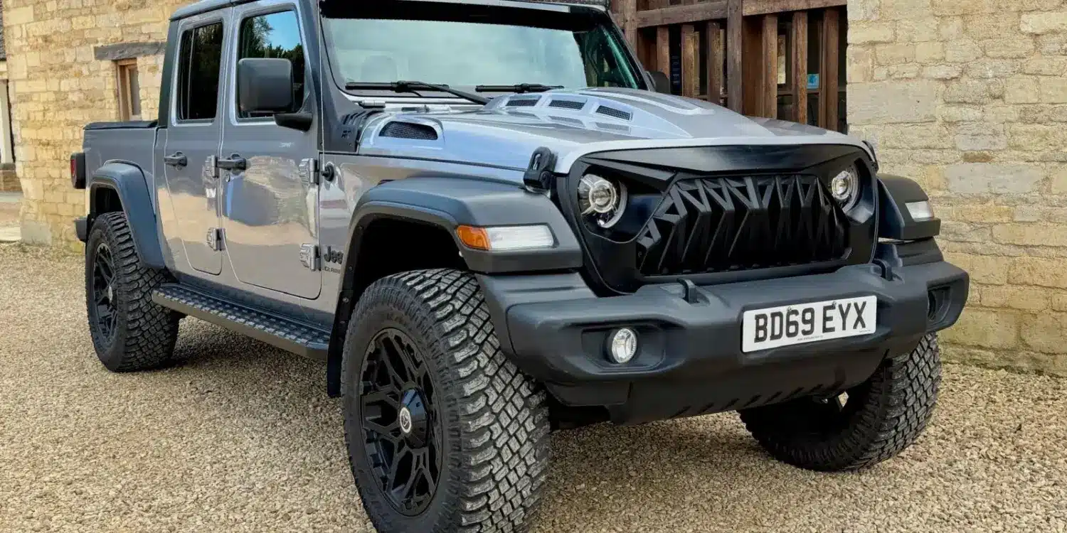 Jeep Gladiator 3.6 V6 Doppelkabine, Grau Metallic, schwarze Akzente, LED-Lichtleiste und Spezial-Front, vor einer Steinmauer
