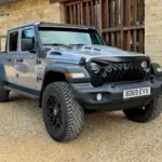 Jeep Gladiator 3.6 V6 Doppelkabine, Grau Metallic, schwarze Akzente, LED-Lichtleiste und Spezial-Front, vor einer Steinmauer