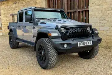 Jeep Gladiator 3.6 V6 Doppelkabine, Grau Metallic, schwarze Akzente, LED-Lichtleiste und Spezial-Front, vor einer Steinmauer