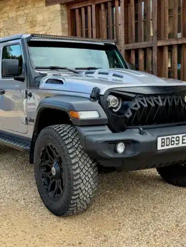 Jeep Gladiator 3.6 V6 Doppelkabine, Grau Metallic, schwarze Akzente, LED-Lichtleiste und Spezial-Front, vor einer Steinmauer