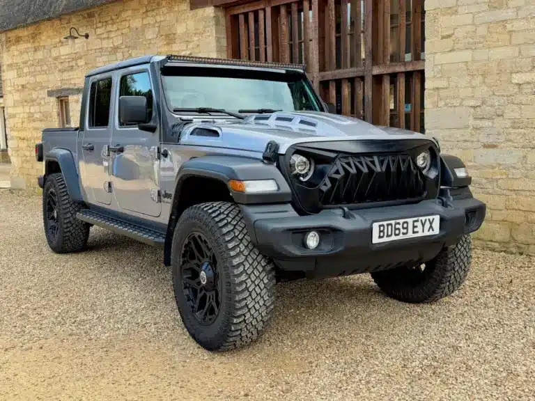 Jeep Gladiator 3.6 V6 Doppelkabine, Grau Metallic, schwarze Akzente, LED-Lichtleiste und Spezial-Front, vor einer Steinmauer