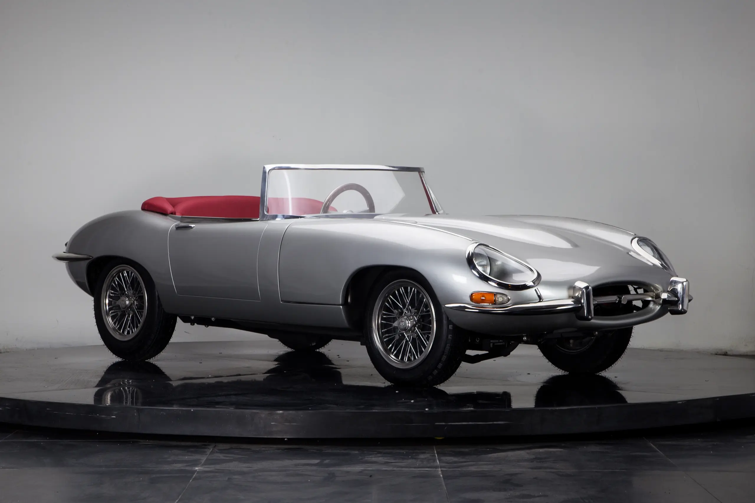 Seitliche Ansicht eines klassischen silbernen Roadsters mit rotem Innenraum vor neutralem Hintergrund.