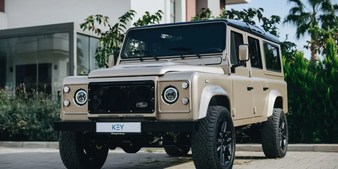 Land Rover Defender 110 V8, Baujahr 1991, beige, vor einem modernen Gebäude geparkt.