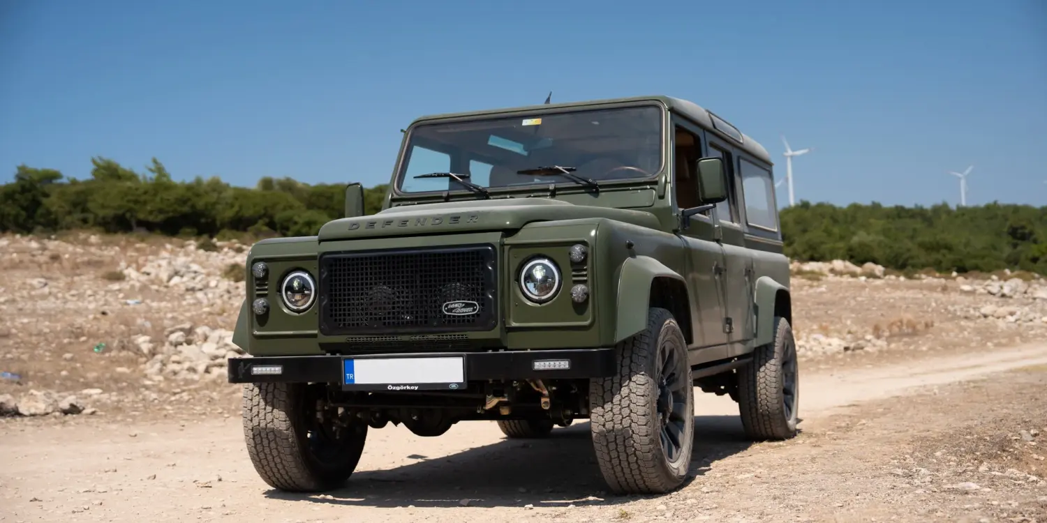 Land Rover Defender 110, Baujahr 1999, in Olivgrün auf Schotterstraße im Gelände vor blauem Himmel.