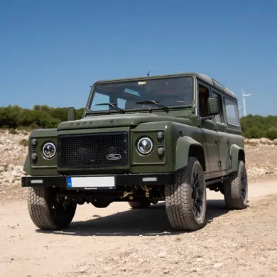Land Rover Defender 110, Baujahr 1999, in Olivgrün auf Schotterstraße im Gelände vor blauem Himmel.