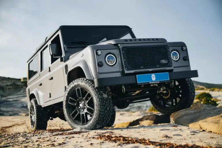 Land Rover Defender 110 in Urban Grey auf sandigem Gelände mit erhöhtem Radstand und Offroad-Bereifung.