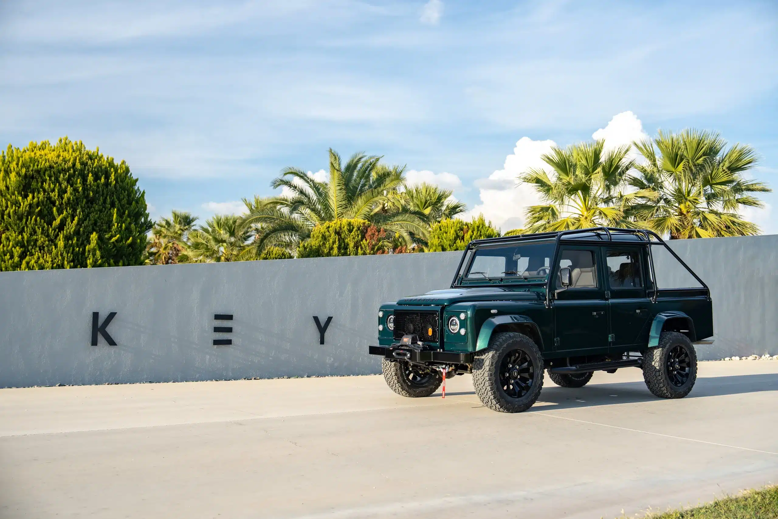 Land Rover Defender 110 von 1994 in Viridian Green Farbe, parkend vor einer weißen Wand mit Palmen und blauem Himmel
